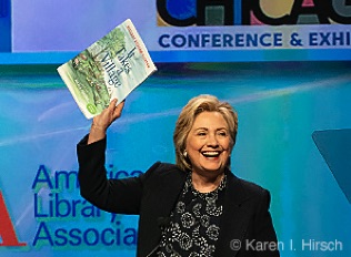 Former Secretary of State Hillary Clinton at the podium raises up her book 